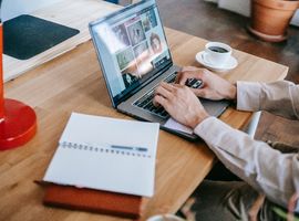 Hombre escribe en un computador en su hogar, acompañado de una taza de café y de un cuaderno de apuntes abierto sobre la mesa.