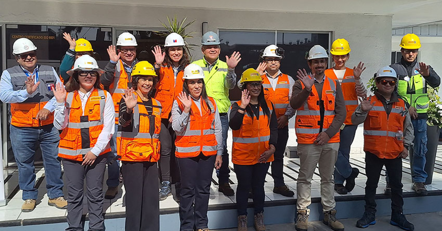 Catorce mujeres y hombres ubicados de píe en dos filas afuera de una empresa, y vestidas con chalecos, zapatos y cascos de seguridad, saludan sonrientes y con sus manos abiertas.